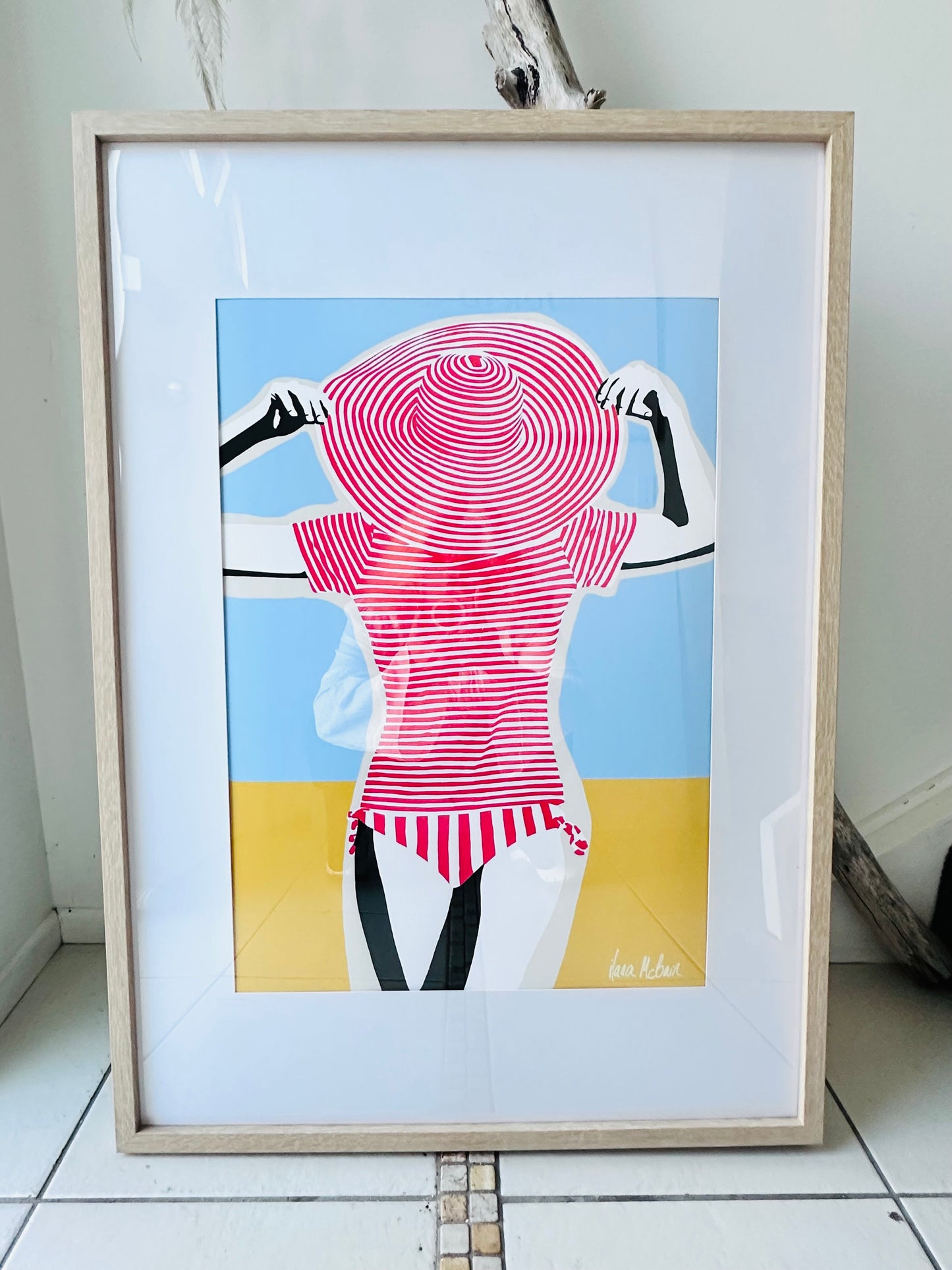 Framed artwork of artistic pop art depiction of a woman wearing large red striped hat and a red and white striped swimsuit against a blue and yellow background.