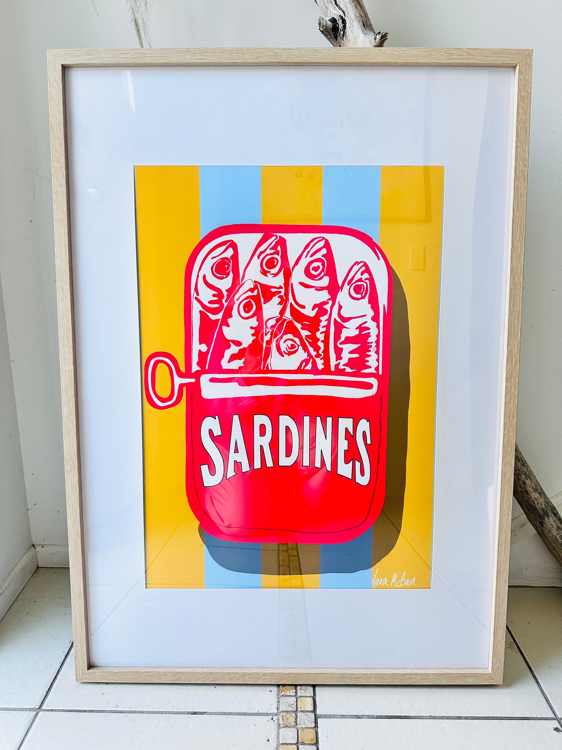 Framed artwork of a red sardine can with fish illustration on a yellow and blue striped background.