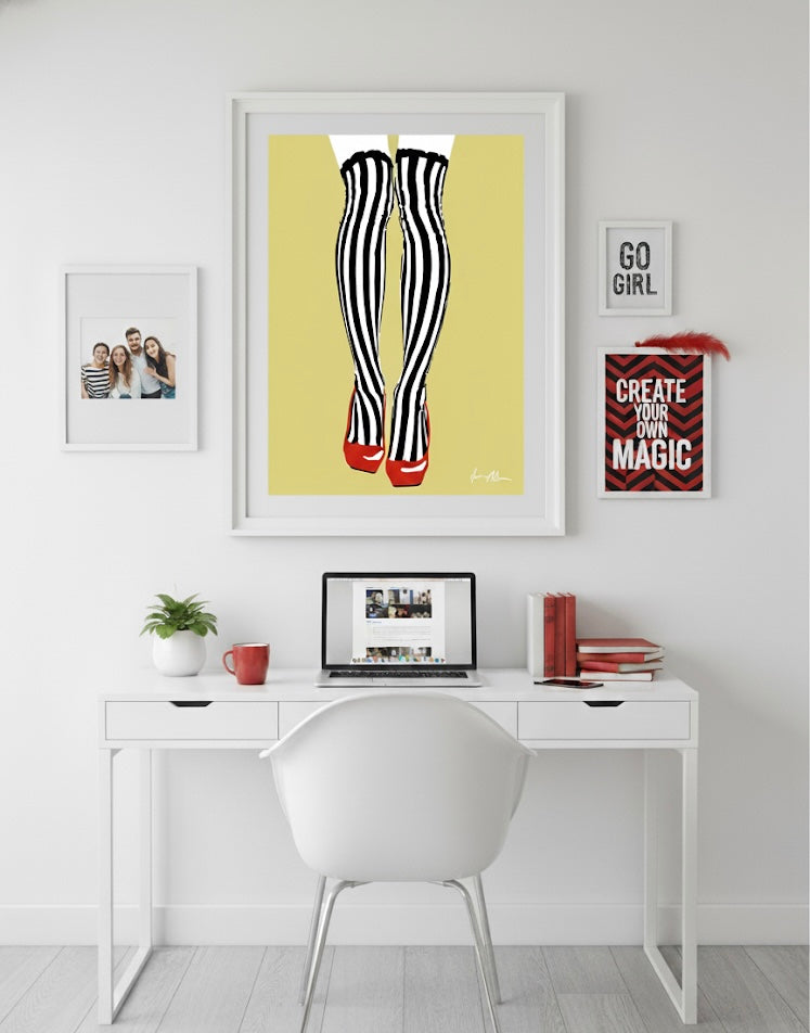 Modern office setup with a desk, chair, and framed artwork of back and white striped tights with red shoes on a golden yellow background on a white wall.